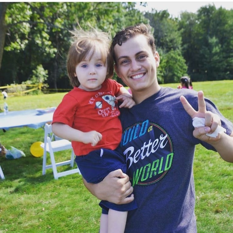 Young man holding a small child at an outdoor park, both smiling at the camera with green grass and picnic setup in background