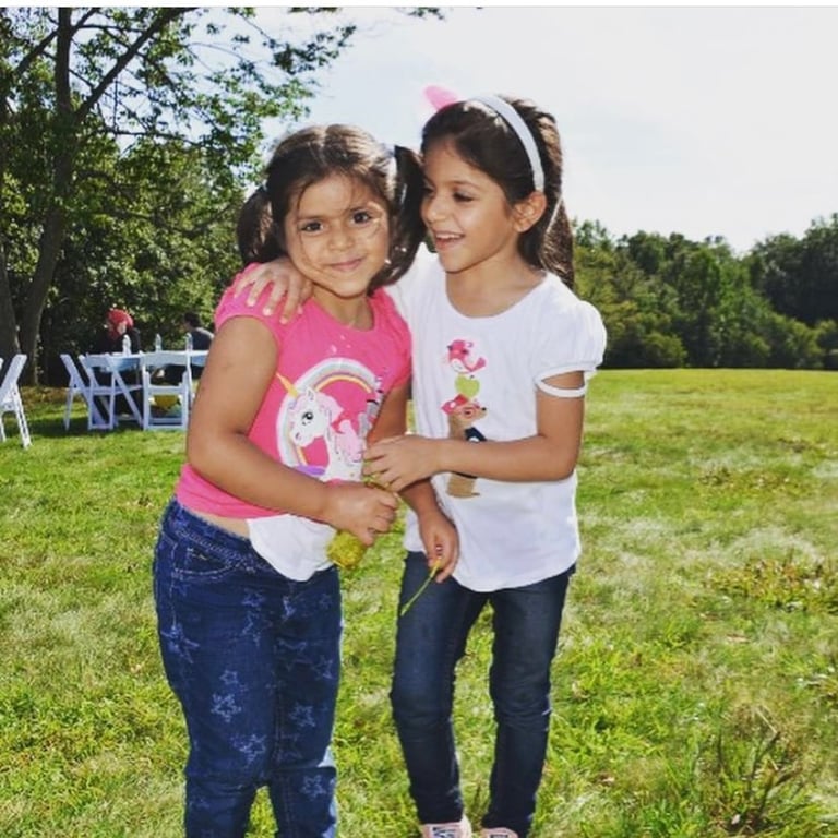Two young girls smiling and posing together outdoors in a grassy field with trees and chairs in the background