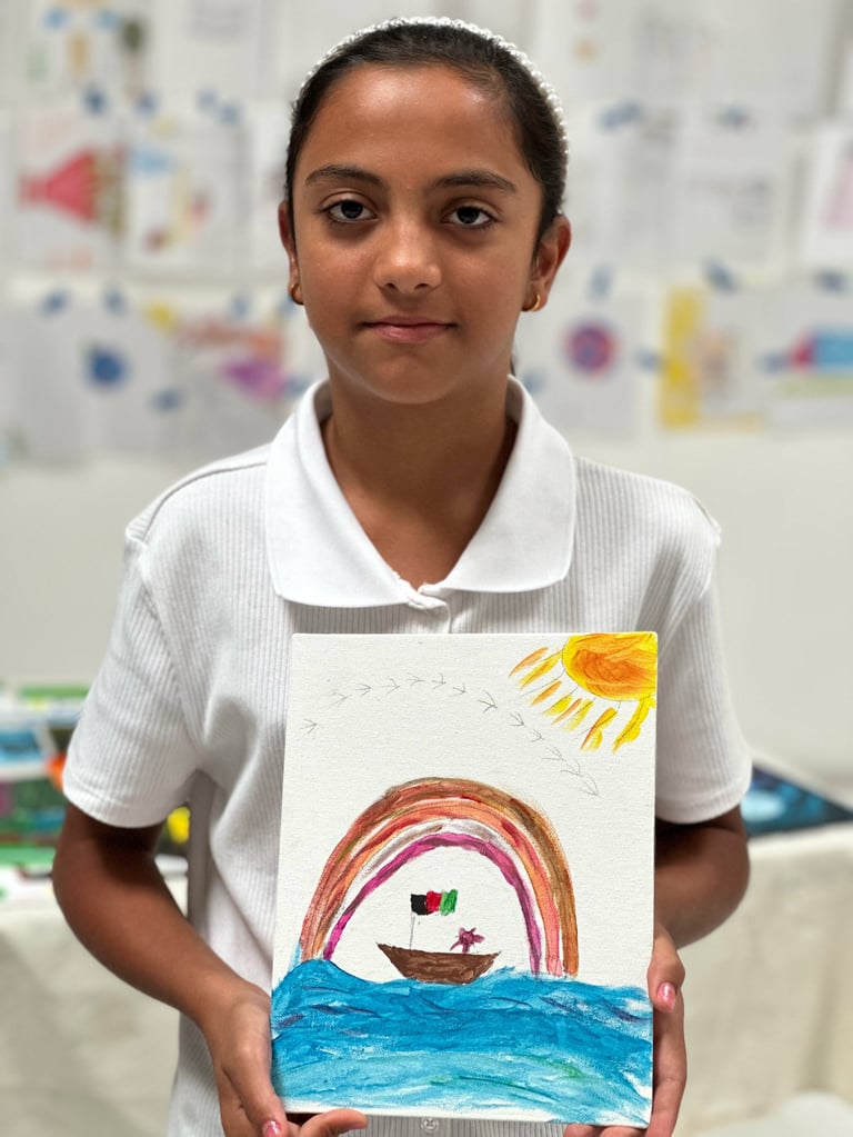 Child holding colorful drawing of sailboat on water with rainbow and sun in classroom