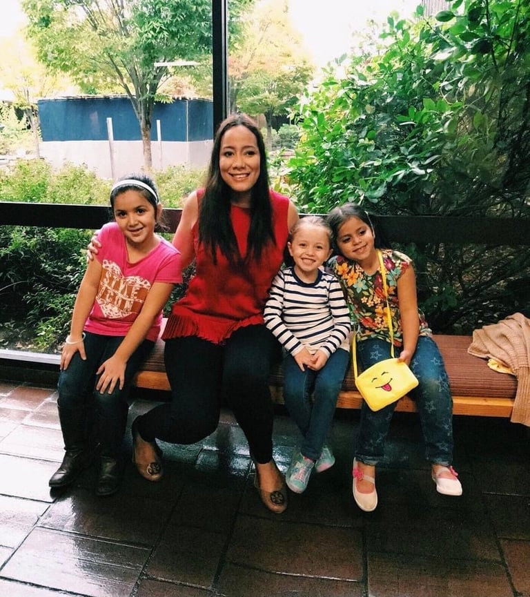 Adult woman with three young girls sitting together on a bench outdoors with greenery and windows in background