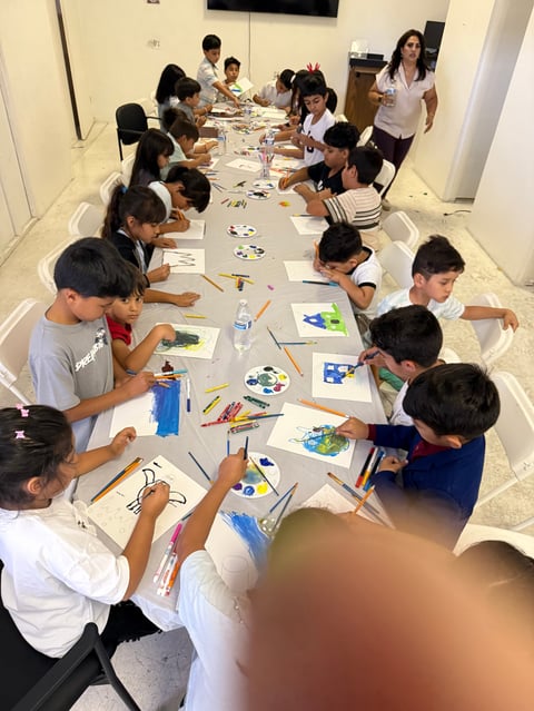 Children and adults seated at a long table doing an art and craft activity with paintbrushes, markers, and colorful drawings