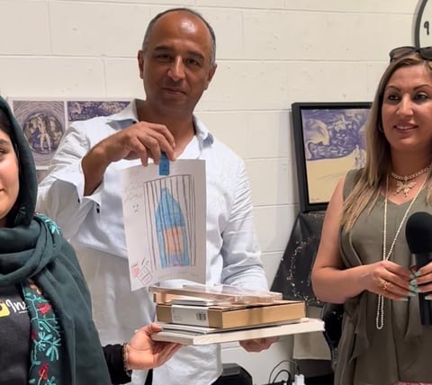 Man in white shirt holds up a drawing while standing between two women in an indoor setting with framed artwork on the wall